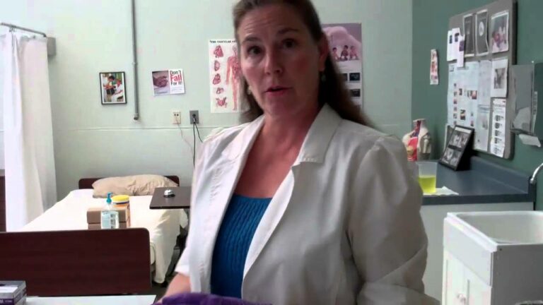 A person in a lab coat stands in a medical room with examination tables and medical posters on the walls.
