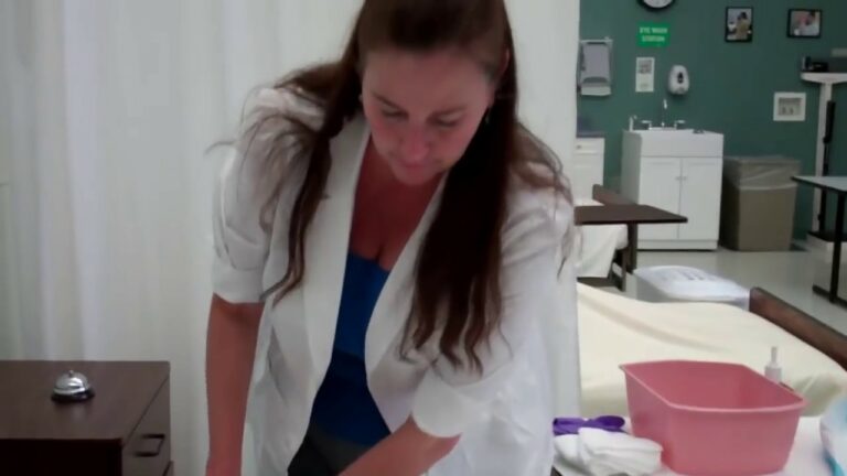 A woman in a white lab coat and blue shirt works in a medical or laboratory setting with equipment and supplies around her.