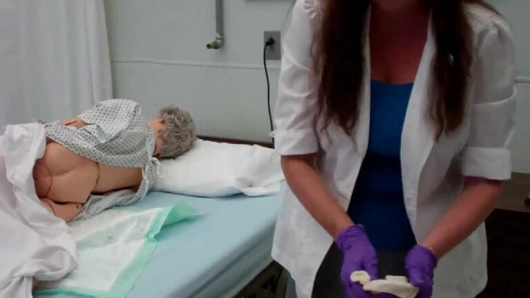 A medical professional wearing gloves stands beside a hospital bed with a patient simulator dummy lying under a blanket.