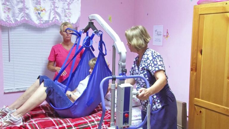Two caregivers assist a person in a blue sling lift above a bed in a pink room.
