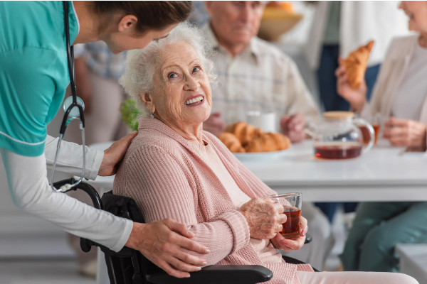 A nurse interacts with a smiling elderly woman in a wheelchair holding a drink, inside a room where other seniors are gathered.