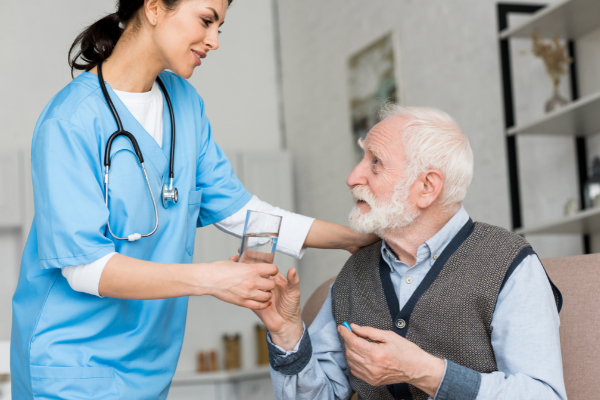 A nurse in blue scrubs gives a glass of water to an elderly man in a vest who is holding a pill.