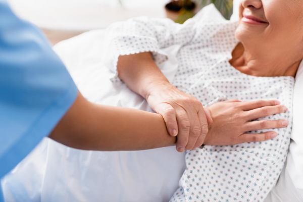 A nurse gently holds the wrist of a smiling patient lying in bed, wearing a hospital gown.