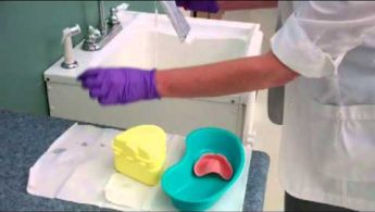 Person in purple gloves pouring liquid into dental mold near a sink, with dentures in a green kidney dish.