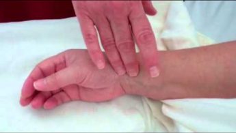 Two hands, one checking the pulse on the wrist of the other, over a white cloth background.