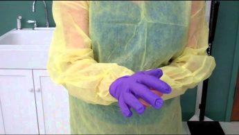 A person wearing a yellow protective gown and purple gloves, standing in front of a white sink in a medical setting.